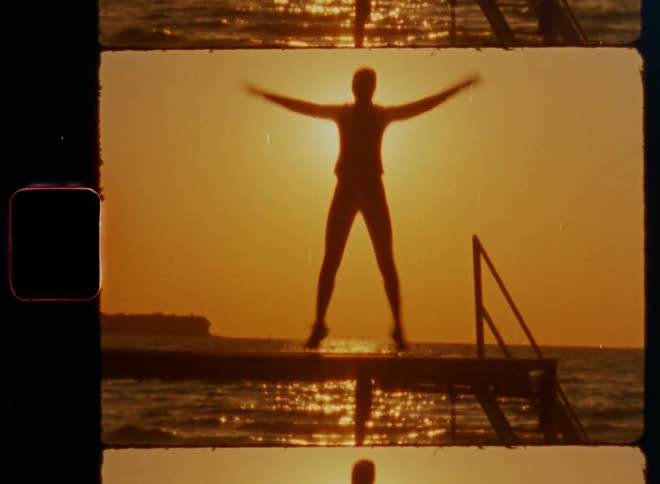 Old Super-8 film footage of a sporty woman exercising working out and doing jumping jacks to stay healthy in the evening sun on a wooden landing stage at a sea port as the sun sets over the ocean at the horizon