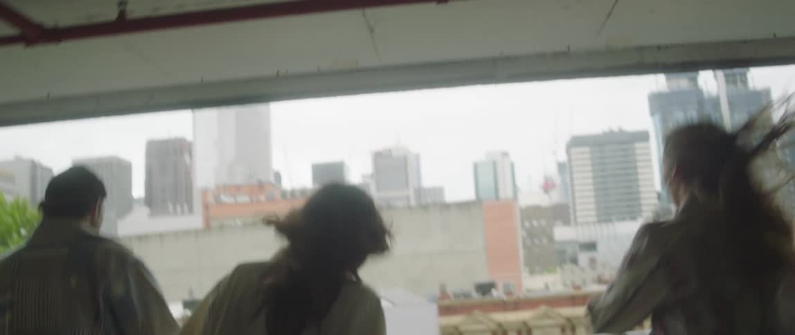 Four people in a parking garage walk to the railing and look out at the city then go to their left