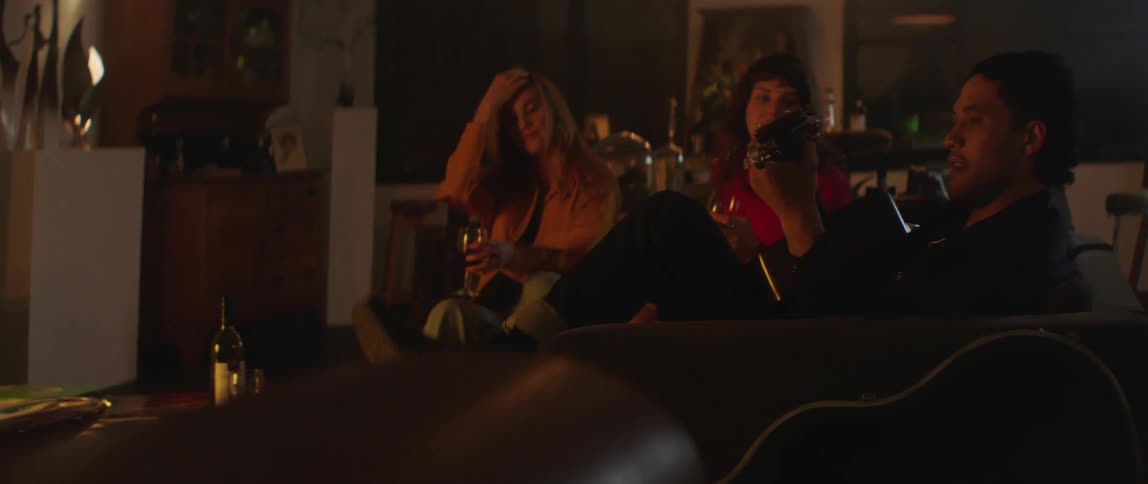 A man sits at home with two women who are drinking wine and he picks up his guitar out of its case and begins go play as they all laugh and smile