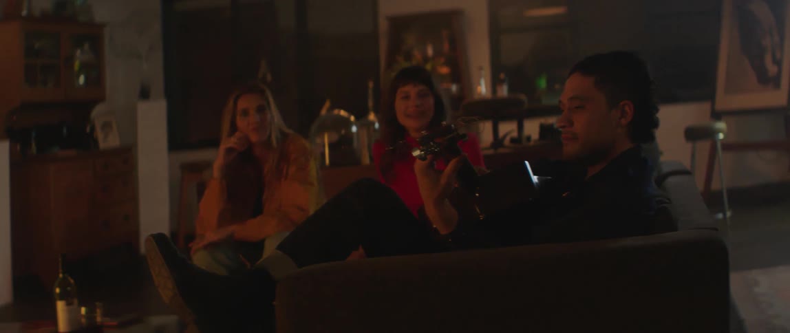 A man sitting on couch in the living room picks up his guitar and starts playing for the two women who are sitting next to him in a social encounter