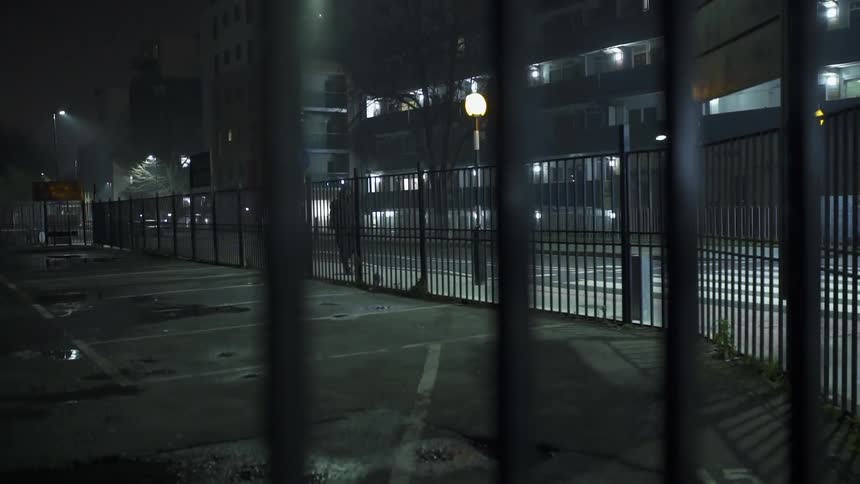 A man wearing dark clothing is walking past a fenced in court at night