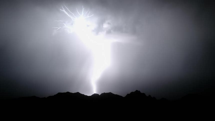 Lightning bolts flash through clouds and strike ground near mountains at night