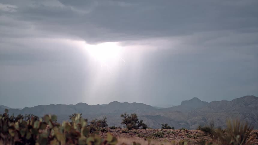 During dusk a lightning strike illuminates the wilderness casting a bright light across the sky near the distant mountains the foreground features sturdy grass growing amidst the sandy terrain