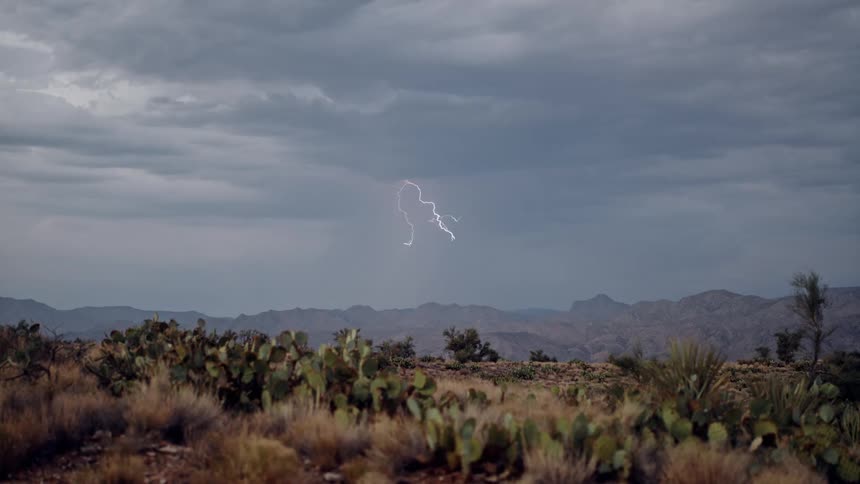 A lightning storm on a cloudy day over a mountain range in the desert with plants on the flatland