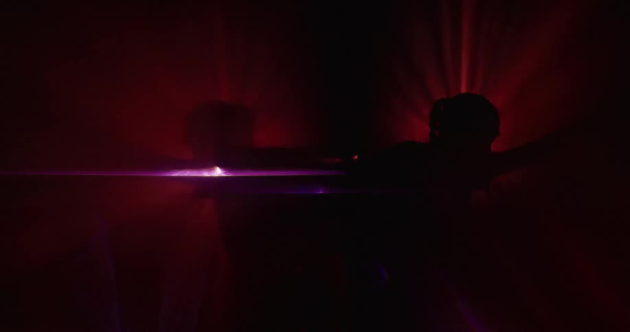 Two mirrored silhouettes moving on a black background with beams of red light and particles in the air