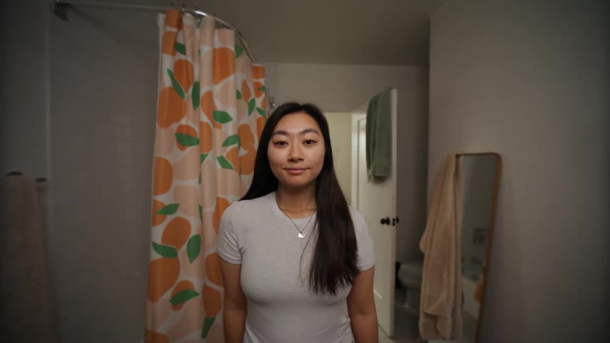 Woman with straight hair gray t shirt and necklace looks in mirror smiling in the bathroom