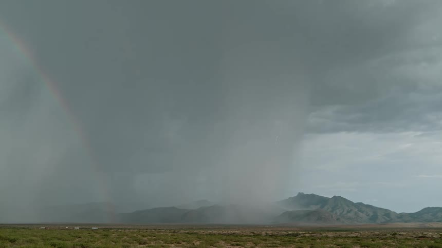 A lightning and rain storm striking down on a mountain range on a flat land with a rainbow across the sky
