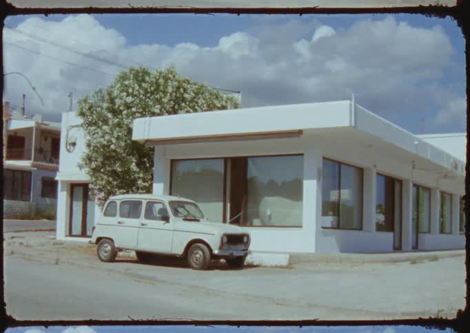An old style car parked outside of a white building on a sidewalk on a bright sunny day