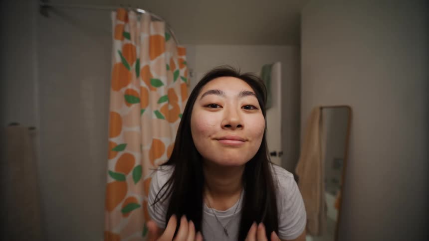 A young woman tends to her hair while looking into a bathroom mirror