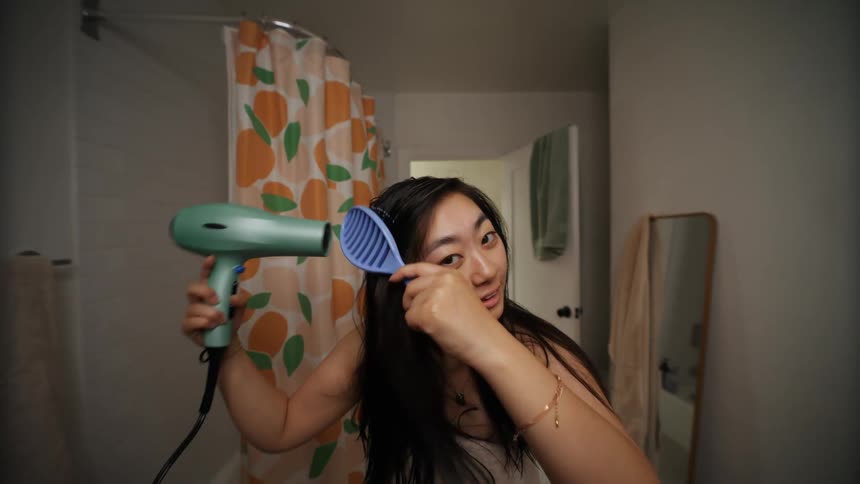 Asian woman indoors in a bathroom blowdrying and brushing her hair