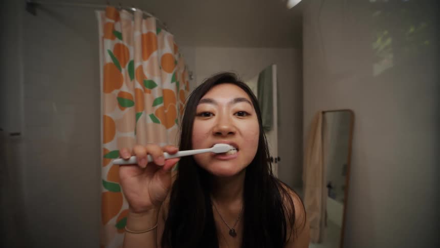A young woman starts by cleaning her mirror then proceeds to brush her teeth before examining her face for any acne in the bathroom
