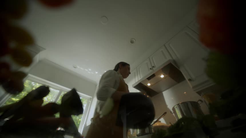 A point of view from a counter top of a woman cooking food in her kitchen and drying her hand on a towel