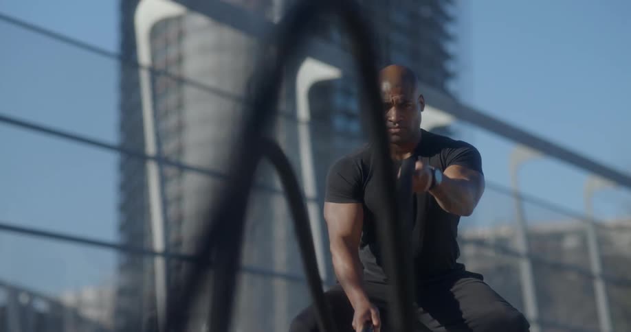 Man squats slightly and whips ropes with buildings in the background