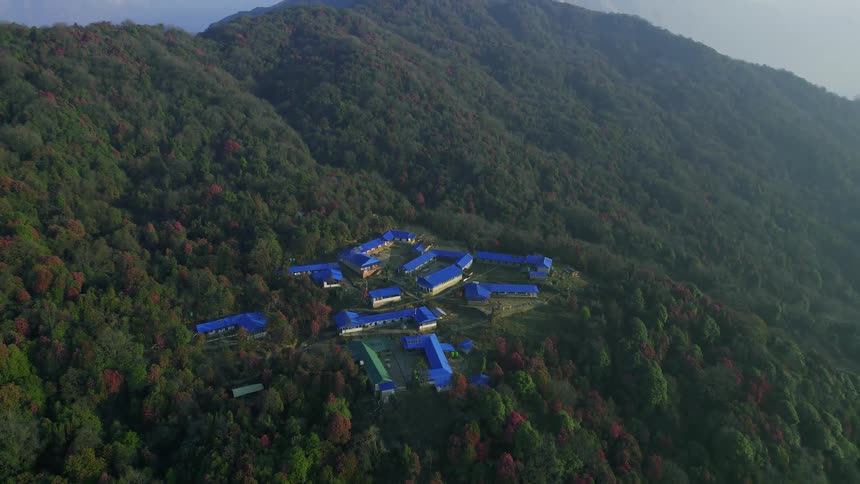 An aerial of houses with blue roofs in the middle of a dense forest on a hillside with tall snow covered mountains in the background