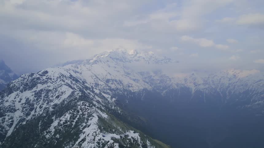 An aerial of snow covered mountain peaks with a mountain summit below on a cloudy day