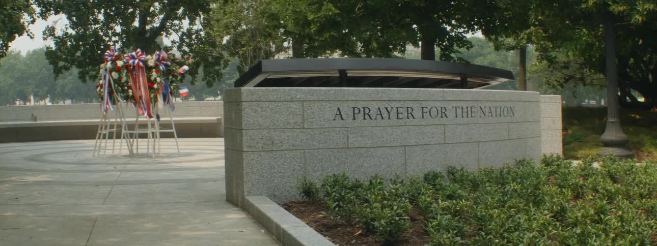 A landmark monument with wreaths and flags surrounded by trees blowing in the wind during the day