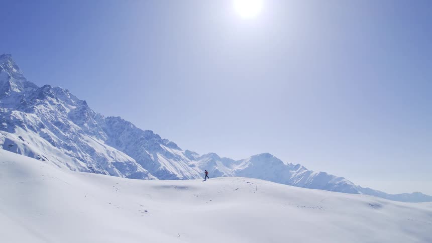 On a sunny day a person strolls on the hill near the mountain where every inch of the land is covered in glistening snow