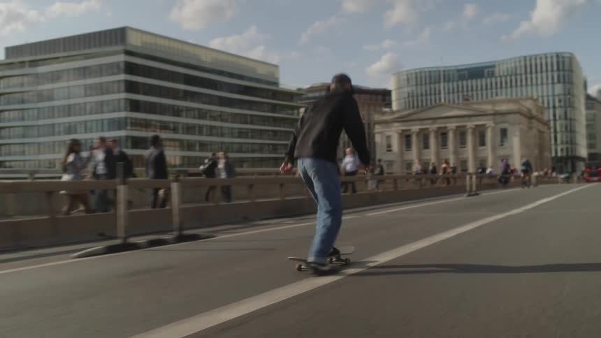 A man wearing a cap riding a skateboard in the middle of the road with people walking on the side walk