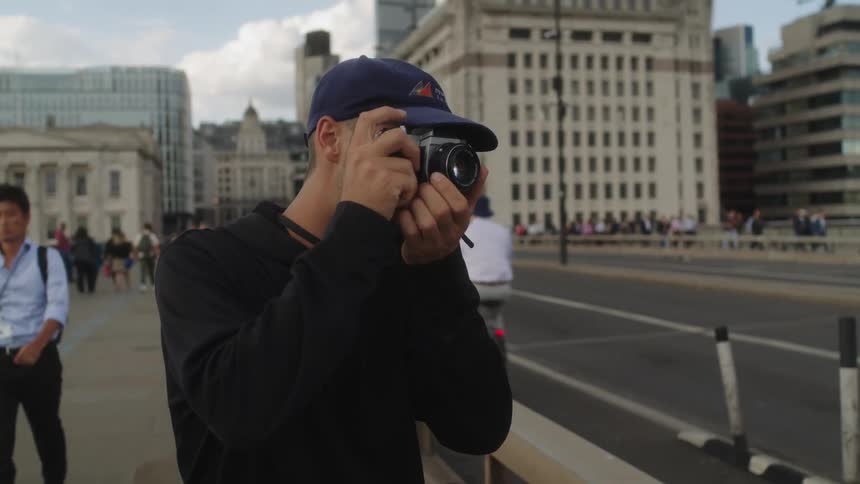 A man wearing a cap stands on the sidewalk capturing a photo with his camera as a biker rides by in the biking lane on a bridge on either side of the bridge people walk by on the walkway while highrise buildings form a striking backdrop