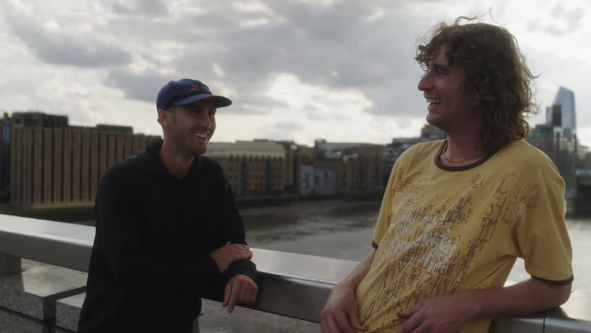 Two men standing leaning on a railing laughing and conversing with a river and city behind them