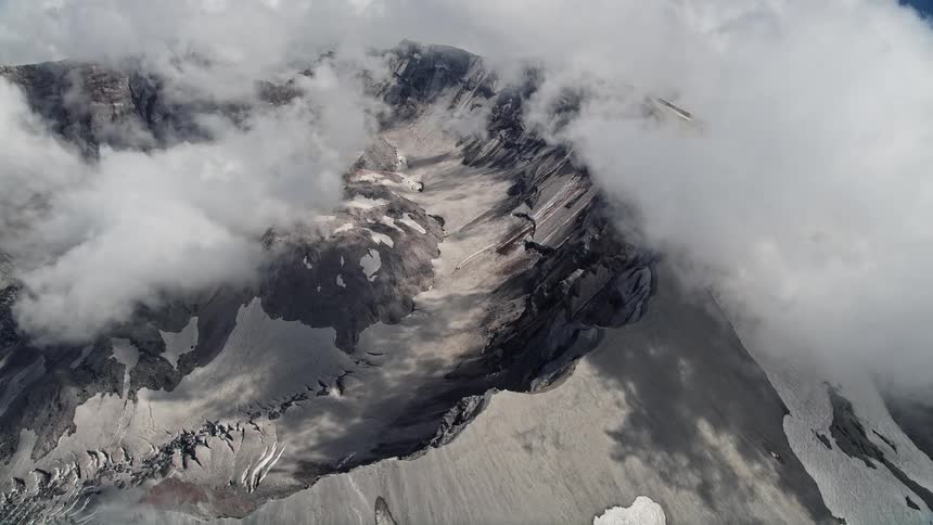 Aerial view pan across and flyby of Mount St Helens crater and clouds in Washington showcasing the majestic snow-covered extinct volcano during daytime