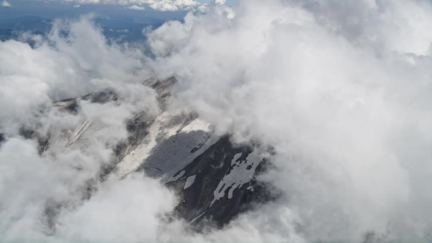 An aerial shot captures the snowy slopes of Mount St Helens emerging from thick clouds in Washington during summer