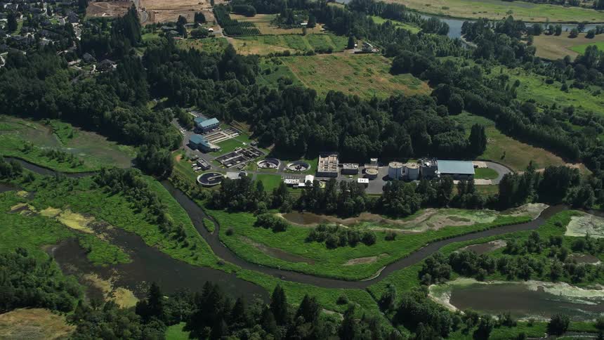 Approaching an industrial water treatment plant nestled beside a river in Ridgefield Washington with lush trees surrounding a facility featuring multiple buildings and storage silos
