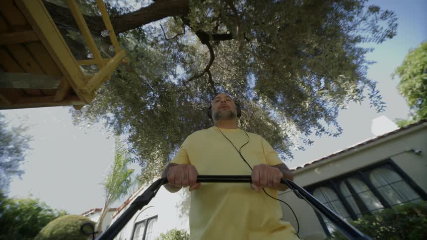 Man wearing headphones pushes mowers across lawn near tree house