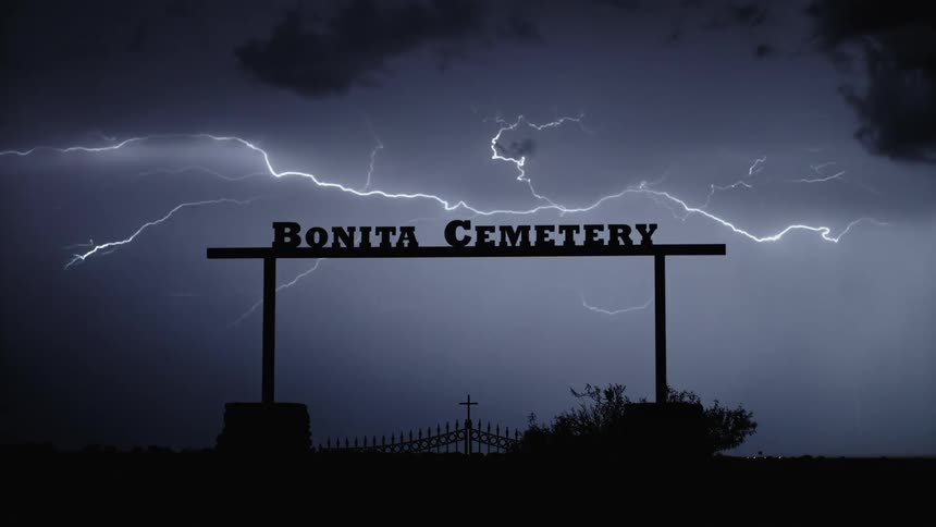 A thunderstorm with lightning lighting up the dark night sky revealing a silhouette of an entrance to a cemetery