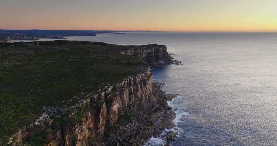 The waves crash against the shore which is shielded by rocks and surrounded by a massive cliff