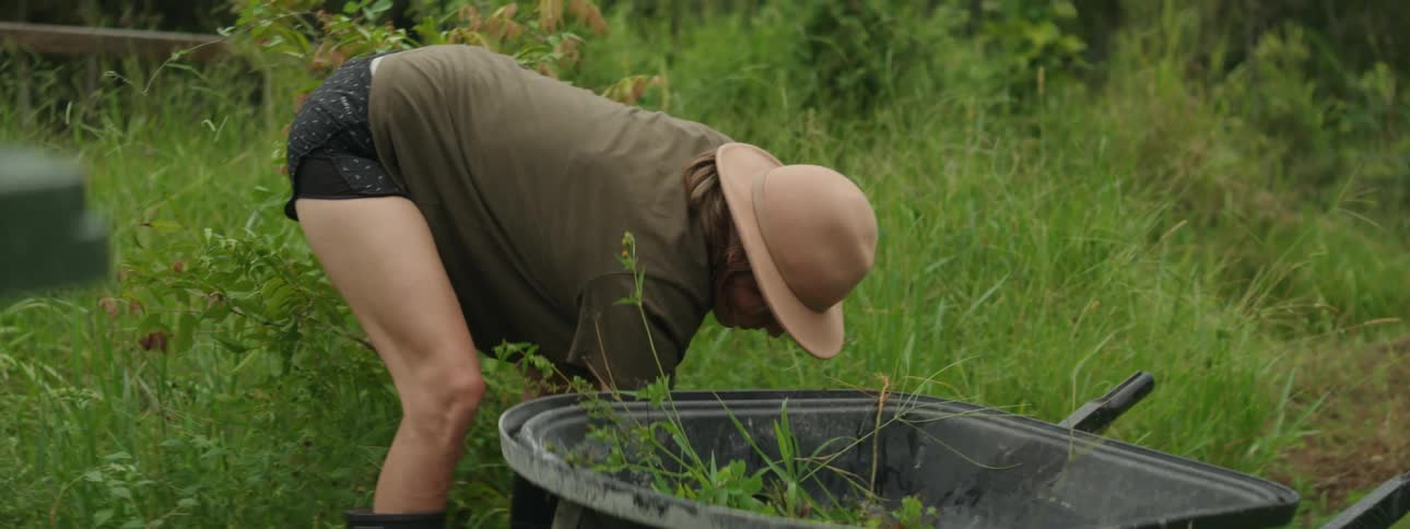 Woman bent over pulling weeds from the ground before placing them in a wheelbarrow next to her