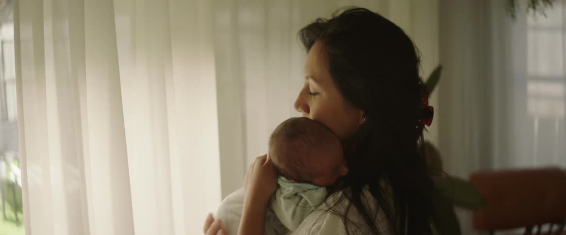 A mother holding her newborn baby standing in front of a bright window in the middle of the day