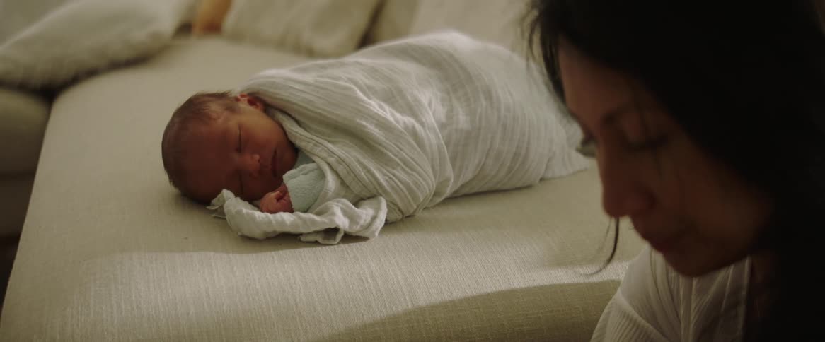 Close up of a woman sitting next to a couch with her baby swaddled in a blanket sleeping as she looks down