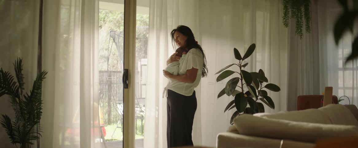 A woman holds an infant child on her shoulder as she shakes him slowly left and right for his comfort inside the living room while standing next to the window