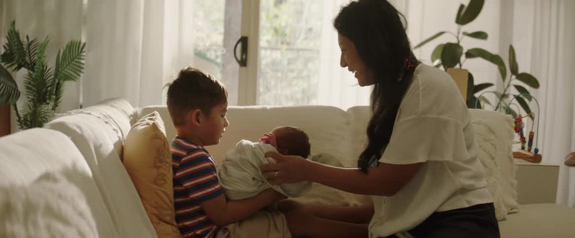 A mother handing her newborn baby to her younger son sitting on a couch in the living room during the day