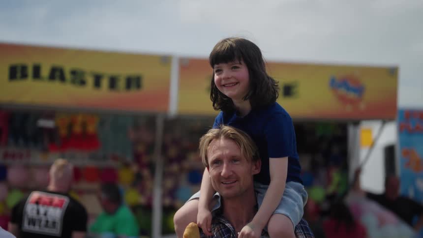 A young girl sitting on her fathers shoulders as they stand in front of a fair booth outdoors on a sunny day smiling laughing and walking forward