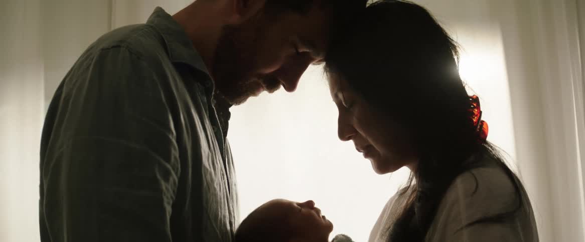 A couple of a man and a woman with their heads touching and eyes closed while holding a baby in their arms