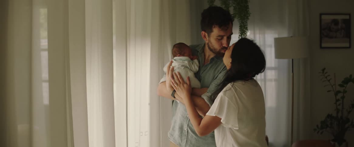 A father holding his newborn baby as his wife walks over and they kiss each other