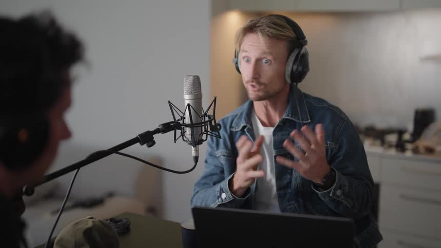 A man wearing headphones and talking into a microphone having a conversation with another man sitting across from him