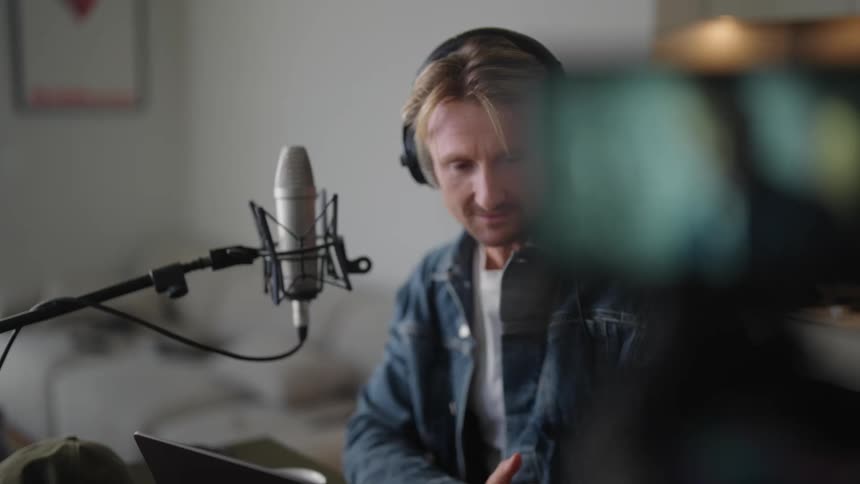 Man sitting in a room in front of a microphone while wearing headphones as he talks and the camera records video