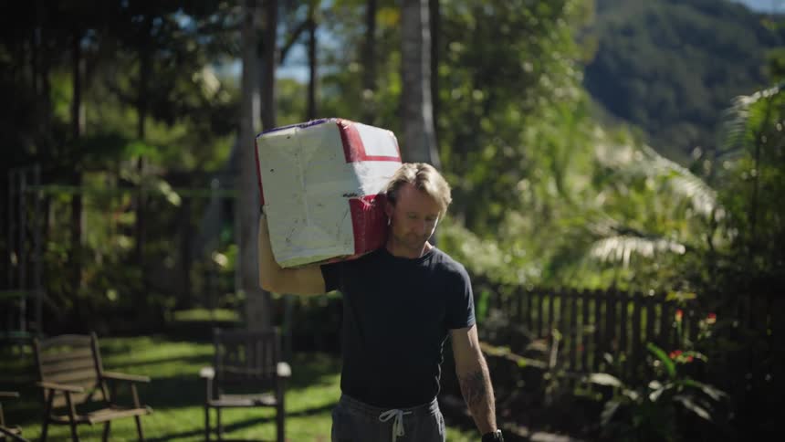 A man carrying a box on his shoulders as he walks forward in a place with a lot of vegetation and trees