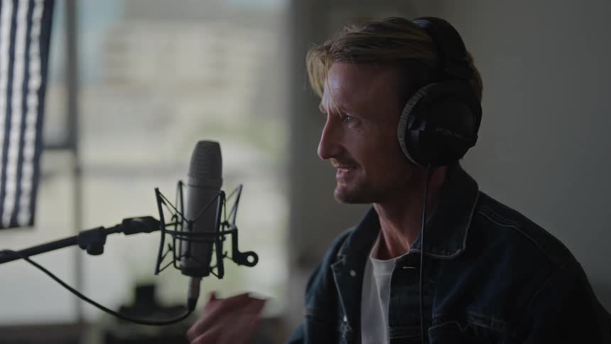 A man wearing headphones and speaking into a microphone while gesturing with his hands indoors during the day