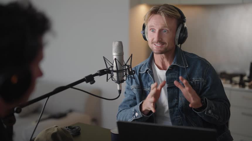 A man wearing headphones talking into a microphone while conversing with a man sitting in front of him in his home in the middle of the day