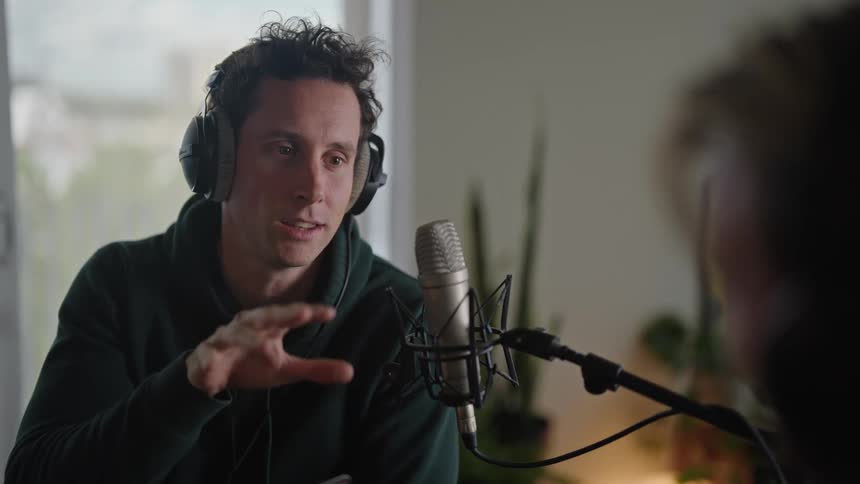 A man with headphones talking into a microphone with a person in front of him while gesturing with his hand