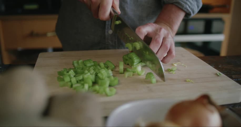 Close up of a man using a knife to cut celery on a cutting board in his kitchen during the day