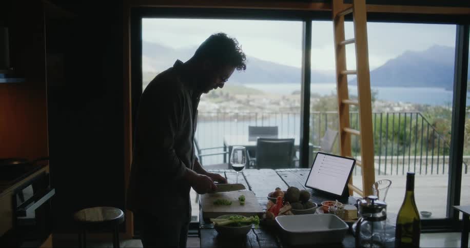 A man stands slicing a cucumber with a knife and a landscape outside his window shows mountains