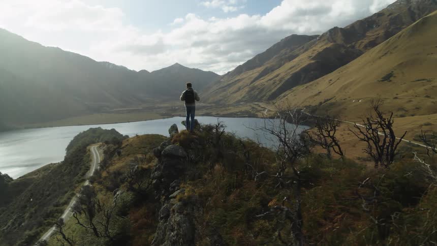 A man stands on the hill in the middle of a lake and looks at the view including the surrounding mountain range