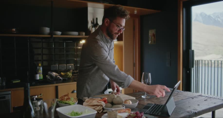 Man standing in his kitchen preparing food while looking at a tablet in front of him