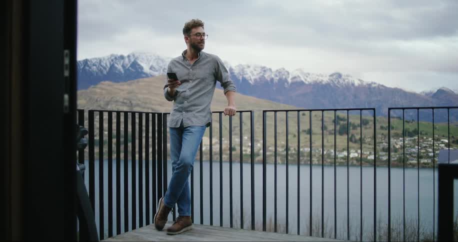 A man standing outside leaning against a black rail while looking at his cellphone a lake and snow covered mountains behind him