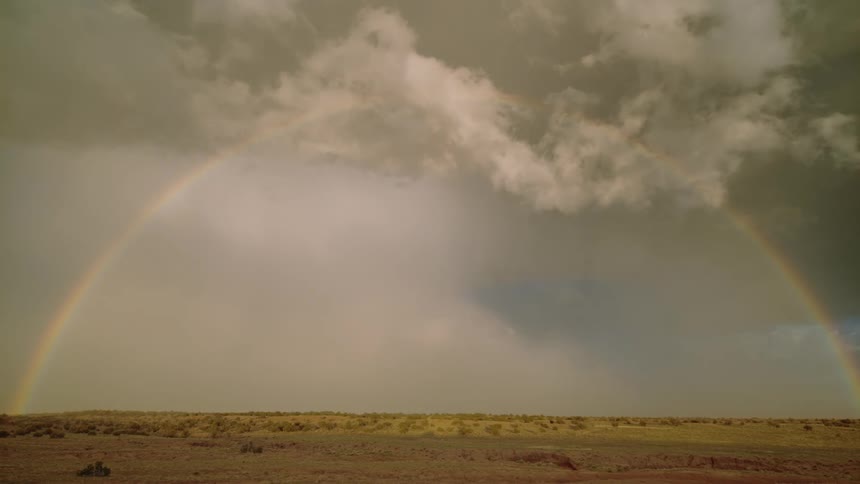 A rainbow cast across the sky over a prairie as lightning flashes in the cloudy sky during the day